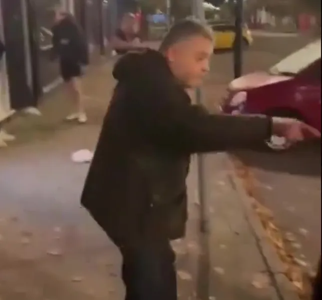Video still of a man appearing to argue with someone on a pavement at night.