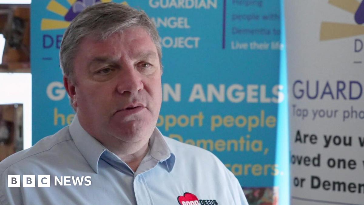 Mark Aldred with grey hair wearing a blue shirt with a Good Deeds Trust logo on it. He stands in front of a Guardian Angel advertising board.