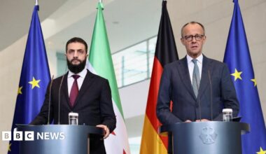 German Chancellor Friedrich Merz and Syrian President Ahmed al-Sharaa wearing suits and standing at podiums, in front of flags, hold a press conference at the Chancellery in Berlin, Germany