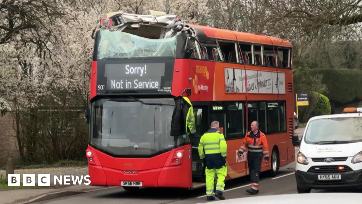 A red and orange double-decker bus is pictured stopped in the road. The upper deck is significantly damaged following a crash with a bridge. A message reading "Sorry! Not in Service" is being shown on the bus