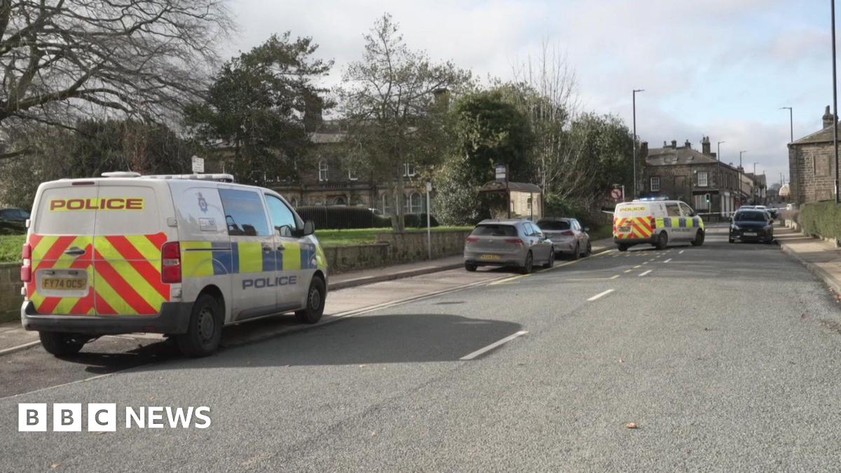 A public street with two police vans and parked cars. A bus stop, trees and other street furniture can be seen.