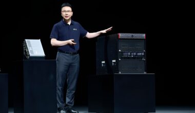 A presenter in a blue polo shirt with a green/yellow logo stands next to a large black server rack on stage, gesturing