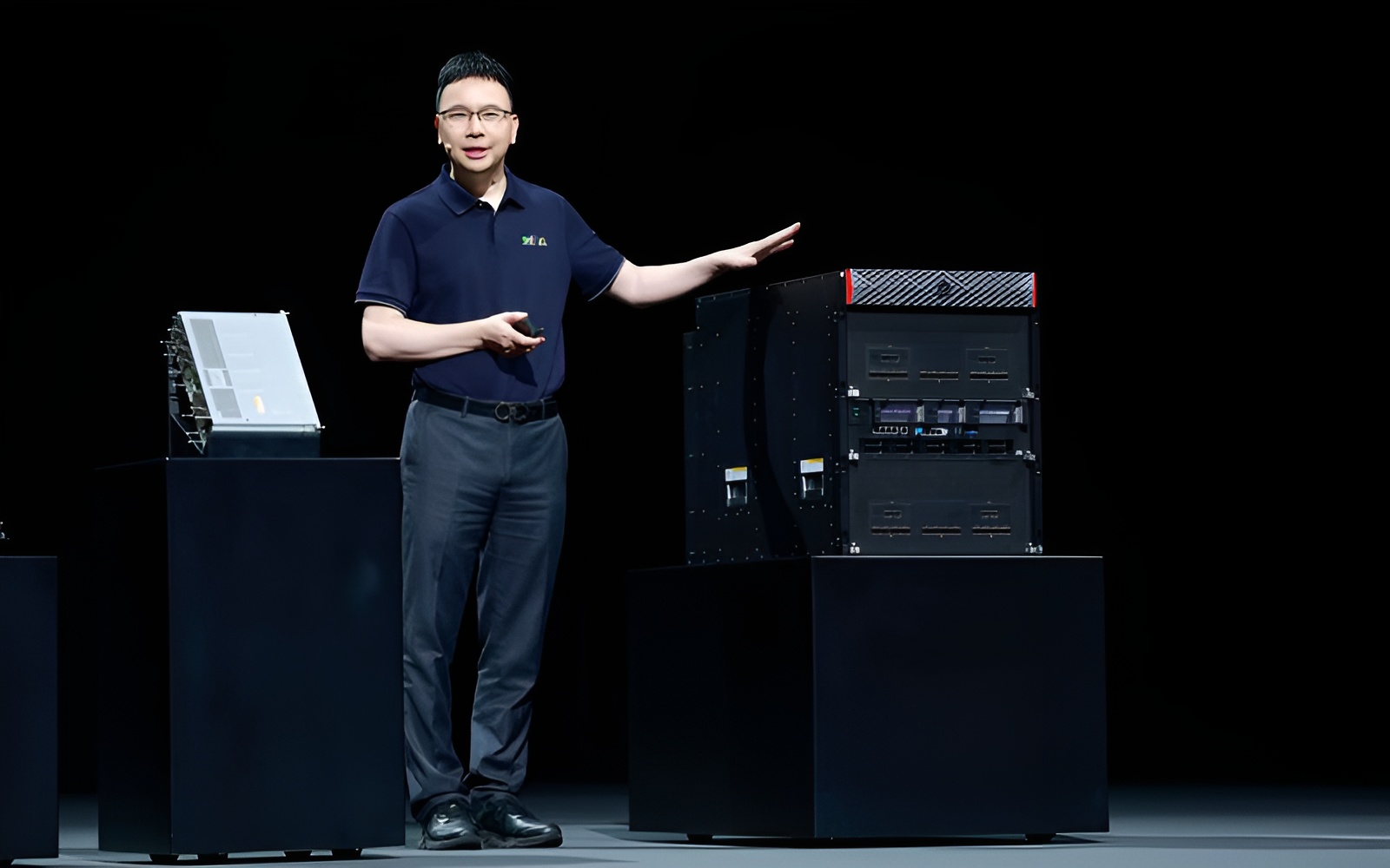 A presenter in a blue polo shirt with a green/yellow logo stands next to a large black server rack on stage, gesturing