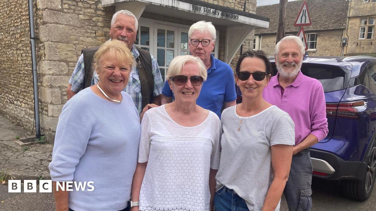 A group of people - three women and three men - stand outside an cold Cotswold stone building with a car outside and a sign on it that says 'the Tolsey Surgery'