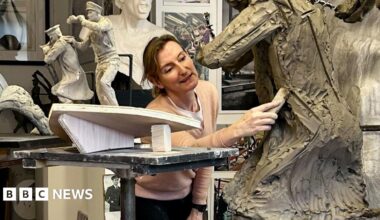 Amy Goodman is pictured in her workshop, using a tool on a statue in progress. Near her are figurines of two men on the prow of a boat, showing how part of the Coastal Forces memorial will look. Photographs of Royal Navy Wrens are positioned in front of her.