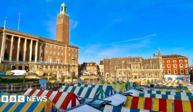 Rows of stalls at Norwich markets in Norwich city centre. The canopies are colourful and striped. In the background is the large imposing building of City Hall, with its tower furthest from the camera. There are blue skies above.