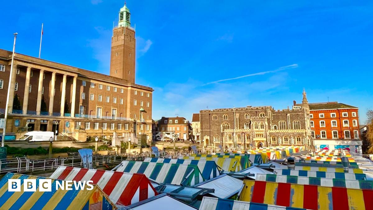 Rows of stalls at Norwich markets in Norwich city centre. The canopies are colourful and striped. In the background is the large imposing building of City Hall, with its tower furthest from the camera. There are blue skies above.