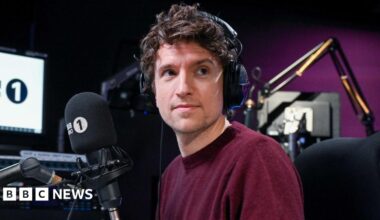 Greg James is wearing a burgundy jumper and is wearing headphones on his head. He is sitting at a desk in front of a microphone with a Radio 1 muff.