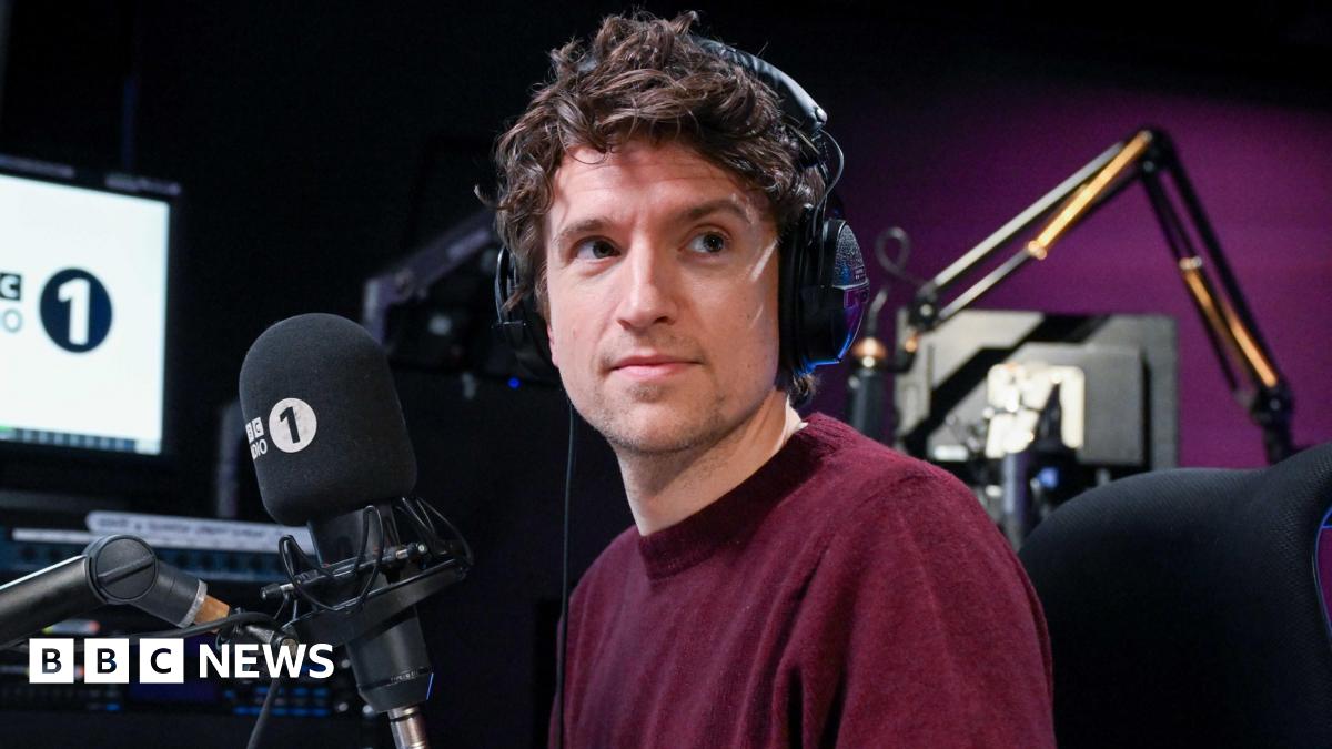 Greg James is wearing a burgundy jumper and is wearing headphones on his head. He is sitting at a desk in front of a microphone with a Radio 1 muff.