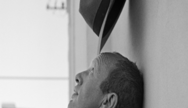 A black and white image of Johnnie Shand Kydd head and neck. It shows his side profile with him leaning his head against a wall while balancing a hat on his head.