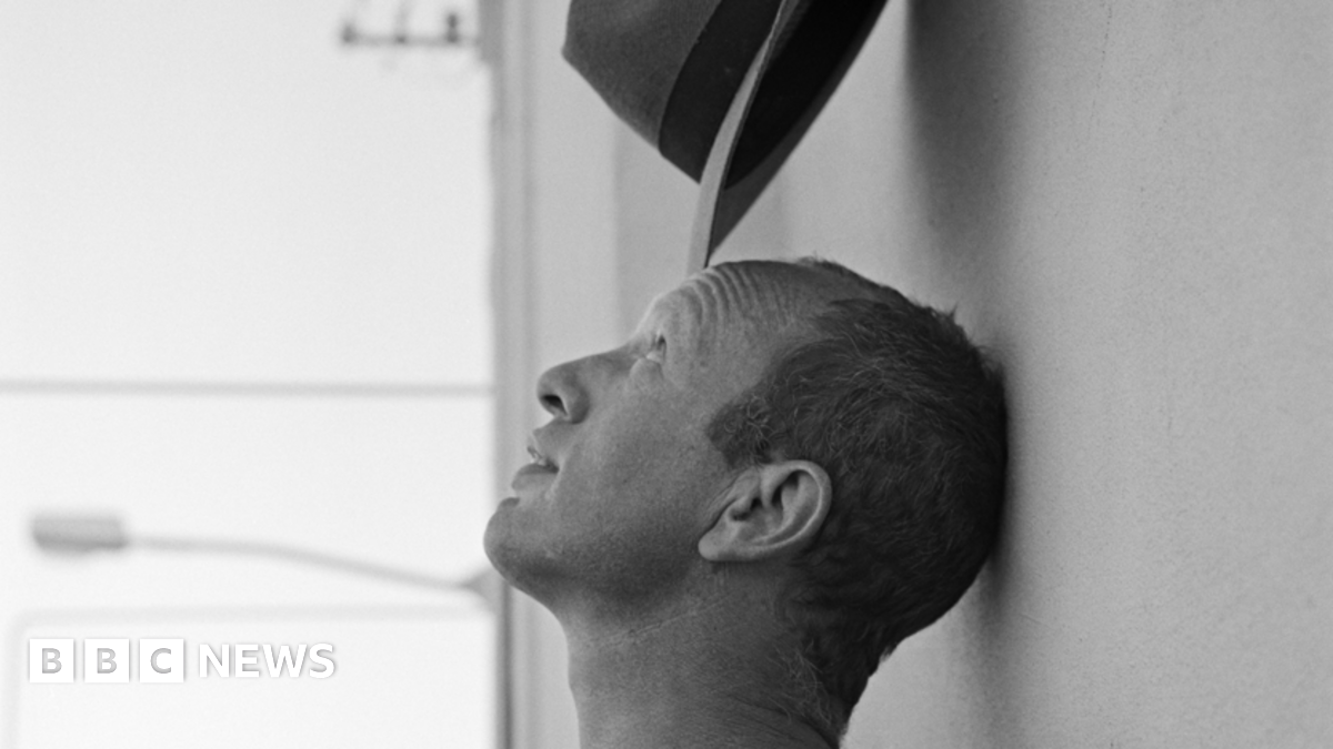 A black and white image of Johnnie Shand Kydd head and neck. It shows his side profile with him leaning his head against a wall while balancing a hat on his head.