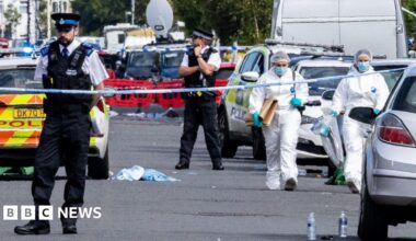Two forensics are walking behind police tape between parked cars. Two police officers are standing behind the cordon.