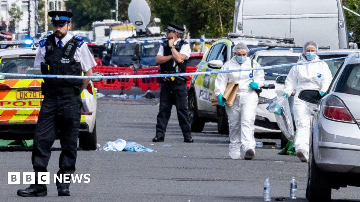 Two forensics are walking behind police tape between parked cars. Two police officers are standing behind the cordon.