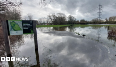 A bench in a flooded open green space area in Datchet. It is sunny.