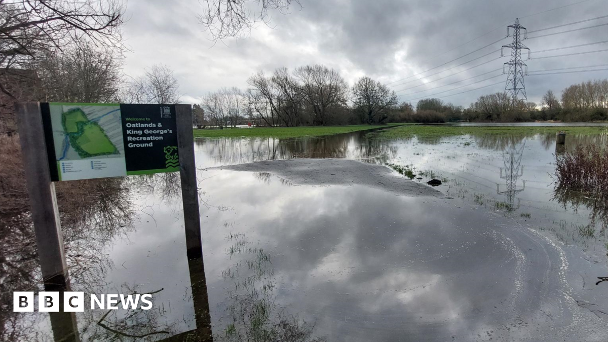 A bench in a flooded open green space area in Datchet. It is sunny.