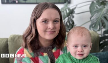 A woman sits with her baby son. She is smiling at the camera. Both are wearing knitted jumpers. A Swiss cheese plant and a green sofa are behind them.