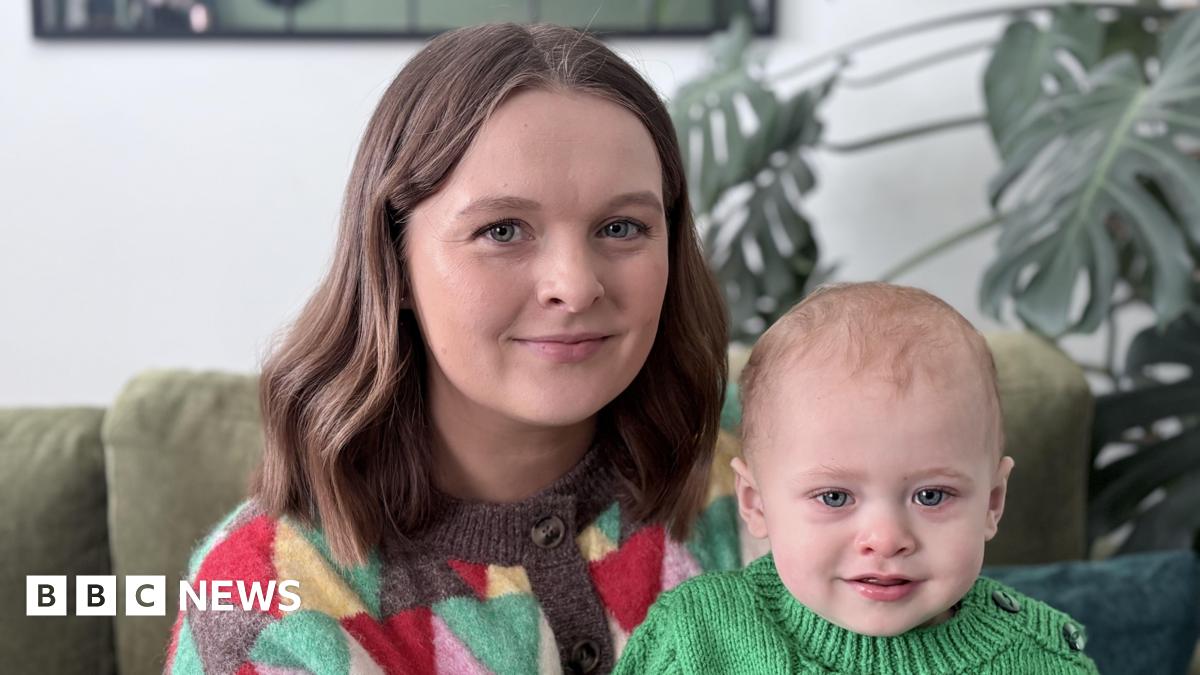 A woman sits with her baby son. She is smiling at the camera. Both are wearing knitted jumpers. A Swiss cheese plant and a green sofa are behind them.
