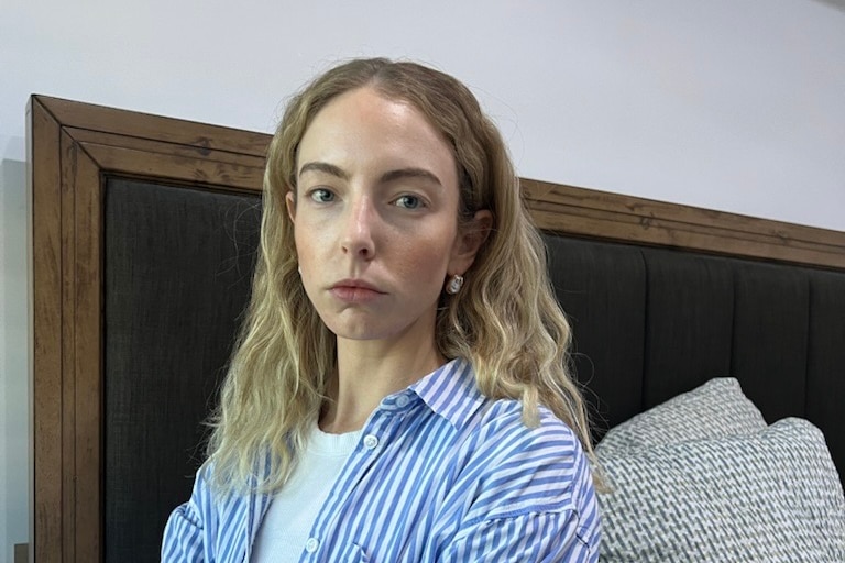 A woman in a blue and white stripey shirt sits on the edge of a bed looking sternly at camera.