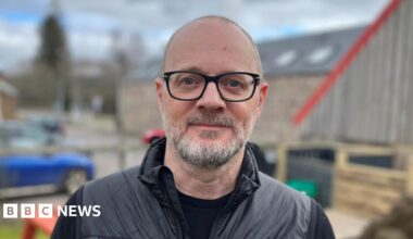 David Watson has grey hair and beard. He is wearing black rimmed glasses and black outdoor clothing. He is standing in a garden area with buildings and a car behind him.