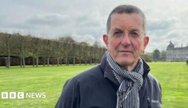 Antony Gormley is standing looking at the camera, wearing a dark grey/navy outdoor jacket and a scarf. In the distance behind him there is a large stately home.
