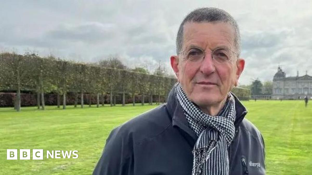 Antony Gormley is standing looking at the camera, wearing a dark grey/navy outdoor jacket and a scarf. In the distance behind him there is a large stately home.
