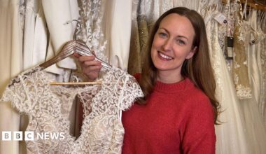 Bronwen Barclay is smiling and holding her wedding gown. Bronwen has long brown hair and is wearing a red crew neck jumper. The top of the dress on a hanger is visible.  It is short sleeved, ivory coloured and made of lace. There are a variety of wedding dresses hanging behind her.