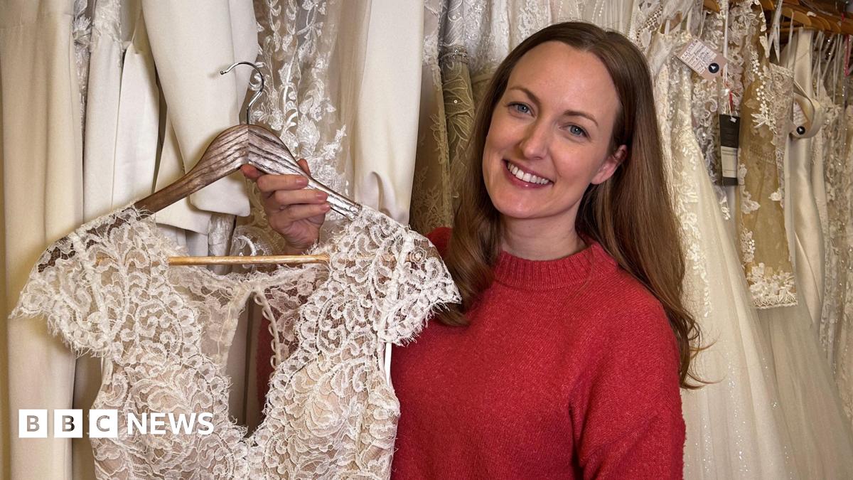 Bronwen Barclay is smiling and holding her wedding gown. Bronwen has long brown hair and is wearing a red crew neck jumper. The top of the dress on a hanger is visible.  It is short sleeved, ivory coloured and made of lace. There are a variety of wedding dresses hanging behind her.
