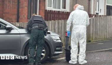 The picture shows a police officer in uniform visible from behind leaning their hands on the bonnet of a car and standing beside another officer dressed in a forensic suit