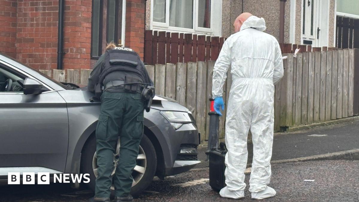 The picture shows a police officer in uniform visible from behind leaning their hands on the bonnet of a car and standing beside another officer dressed in a forensic suit