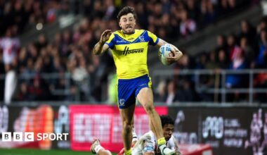 A photo of Matty Ashton running with the ball in his Warrington Wolves kit with the crowd in the background and an opposing player on the floor behind him
