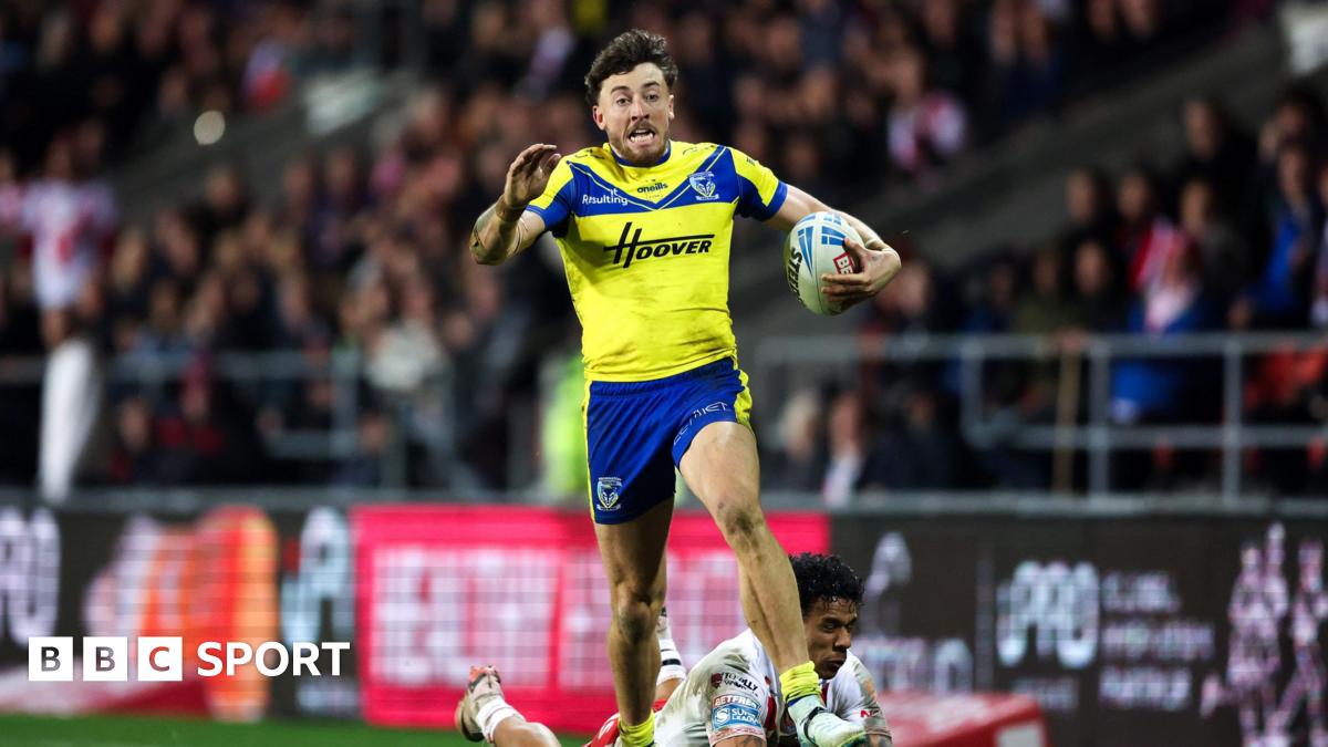 A photo of Matty Ashton running with the ball in his Warrington Wolves kit with the crowd in the background and an opposing player on the floor behind him