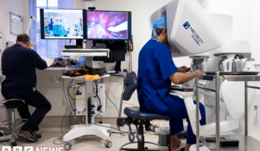 A female consultant in blue scrubs, glasses and face mask operates a machine which carries out robotic hysterectomies.