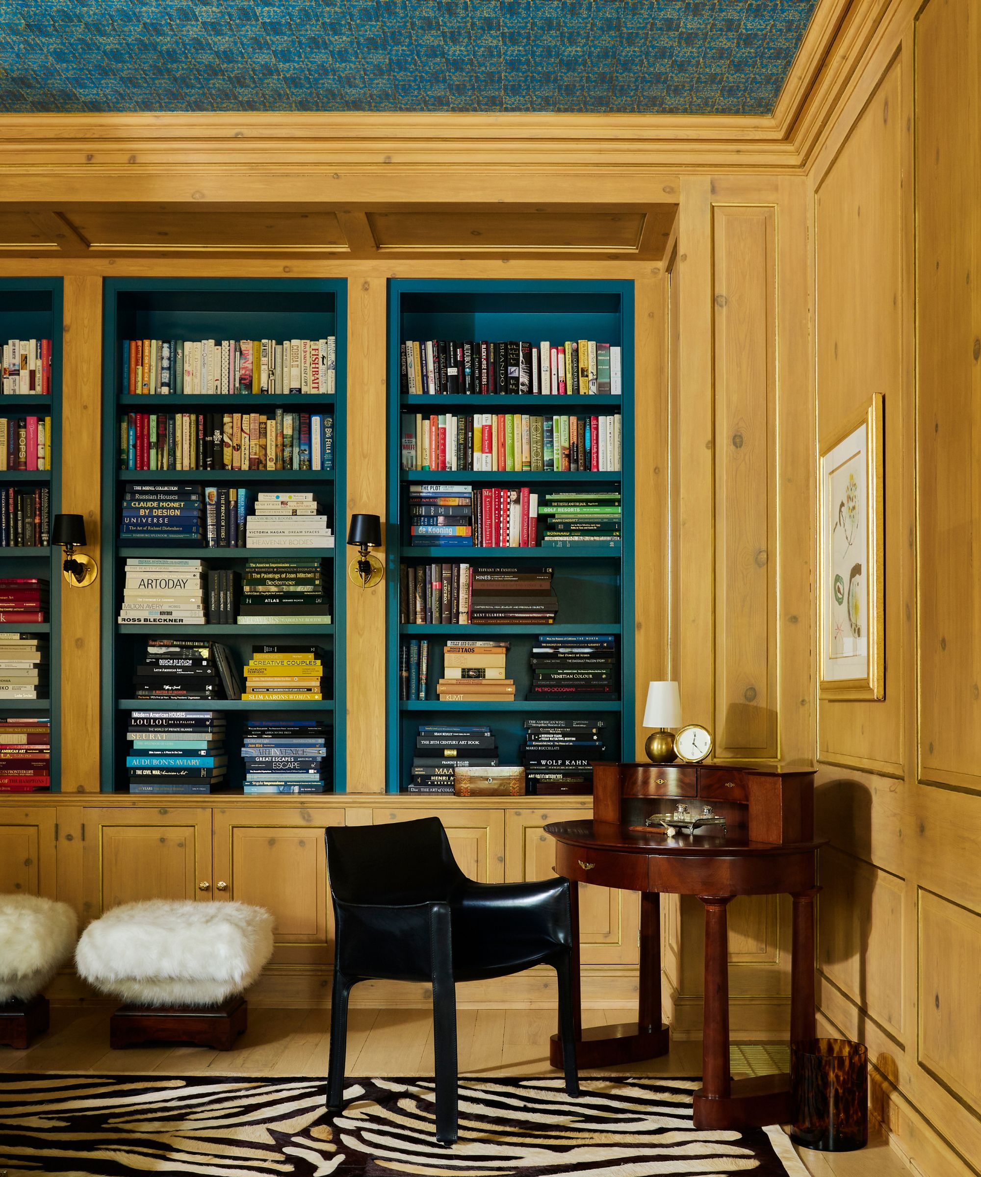 A library room with wood walls, teal built-in bookshelves, a black and white zebra rug, a vintage desk, and teal wallpaper on the ceiling.