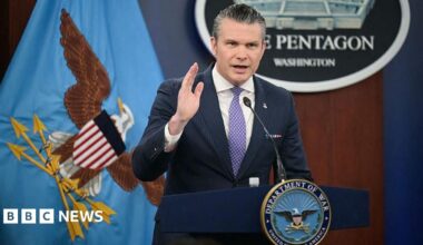 A satellite view of the Strait of Hormuz, with a thin stream of dark blue water flowing around a sharp point of arid land jutting into it.