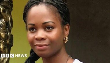 Frances has black braided hair and brown eyes. She is smiling and looking into the distance. She wears gold earrings and a gold necklace.