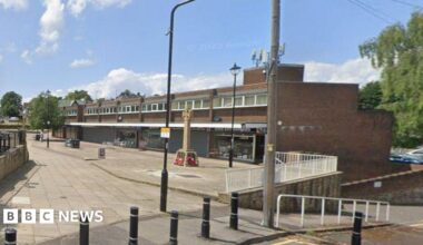 An empty suburban parade of shops. There is a war memorial, lampposts and other street furniture.