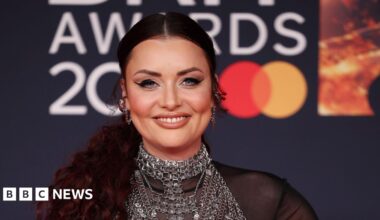 Shona McGarty smiles at the camera while on the red carpet at the Brit Awards. Her hair is tied back and she wears silver chunky earrings and a chain mail style vest over a see-through black blouse.