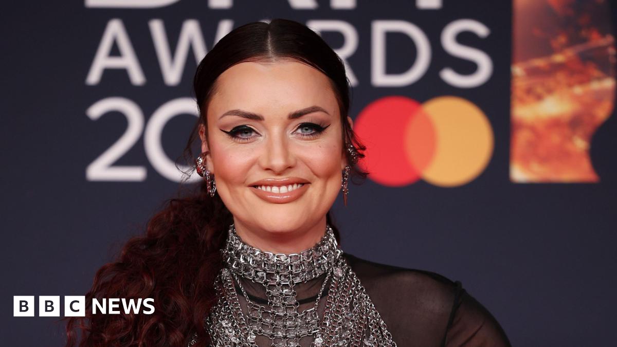 Shona McGarty smiles at the camera while on the red carpet at the Brit Awards. Her hair is tied back and she wears silver chunky earrings and a chain mail style vest over a see-through black blouse.
