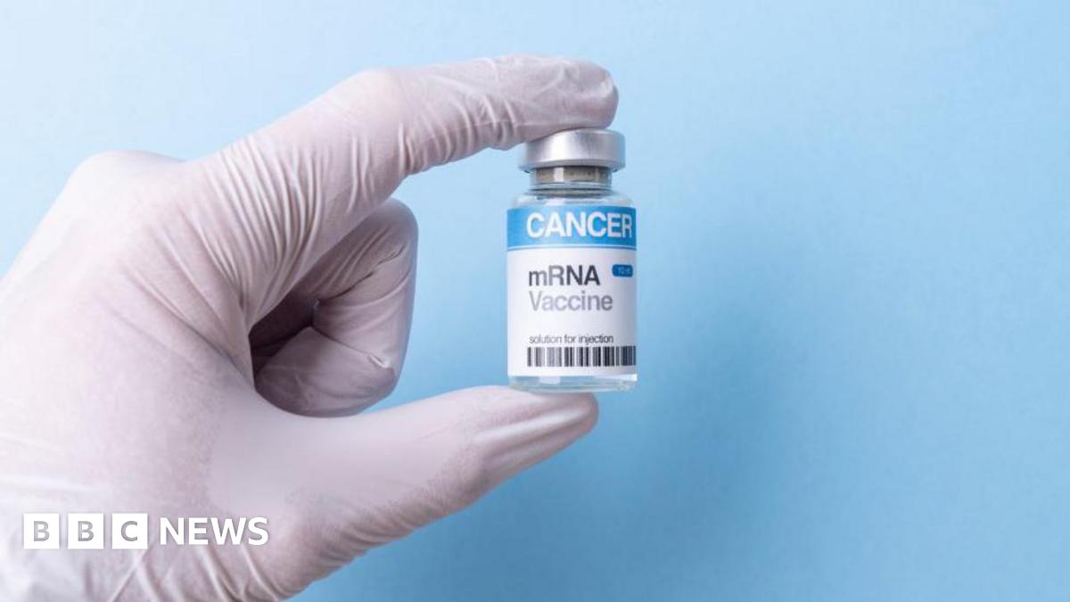 A gloved hand holds up a vial of the mRNA vaccine against a blue background.