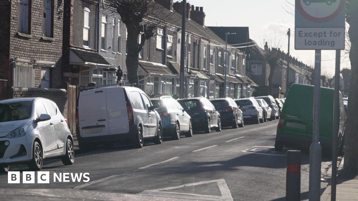 A residential street lined with terraced houses and leafless trees. Cars and vans are parked tightly along both sides of the road. A road sign is visible in the foreground on the right.