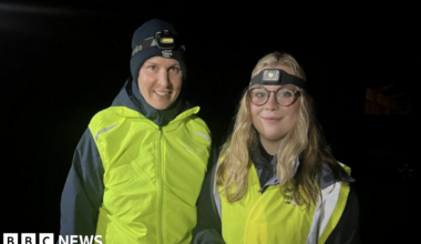 A woman with a black hat and black coat alongside a woman with long blonde hair and glasses. Both are wearing yellow reflective jackets