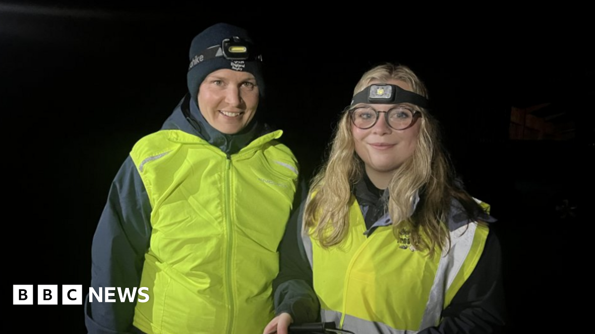 A woman with a black hat and black coat alongside a woman with long blonde hair and glasses. Both are wearing yellow reflective jackets