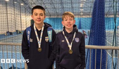 Two young boys in blue hooded tops with medals around their necks. There are indoor tennis courts behind them.