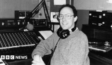 A black and white picture of a man with headphones around his neck wearing a jumper