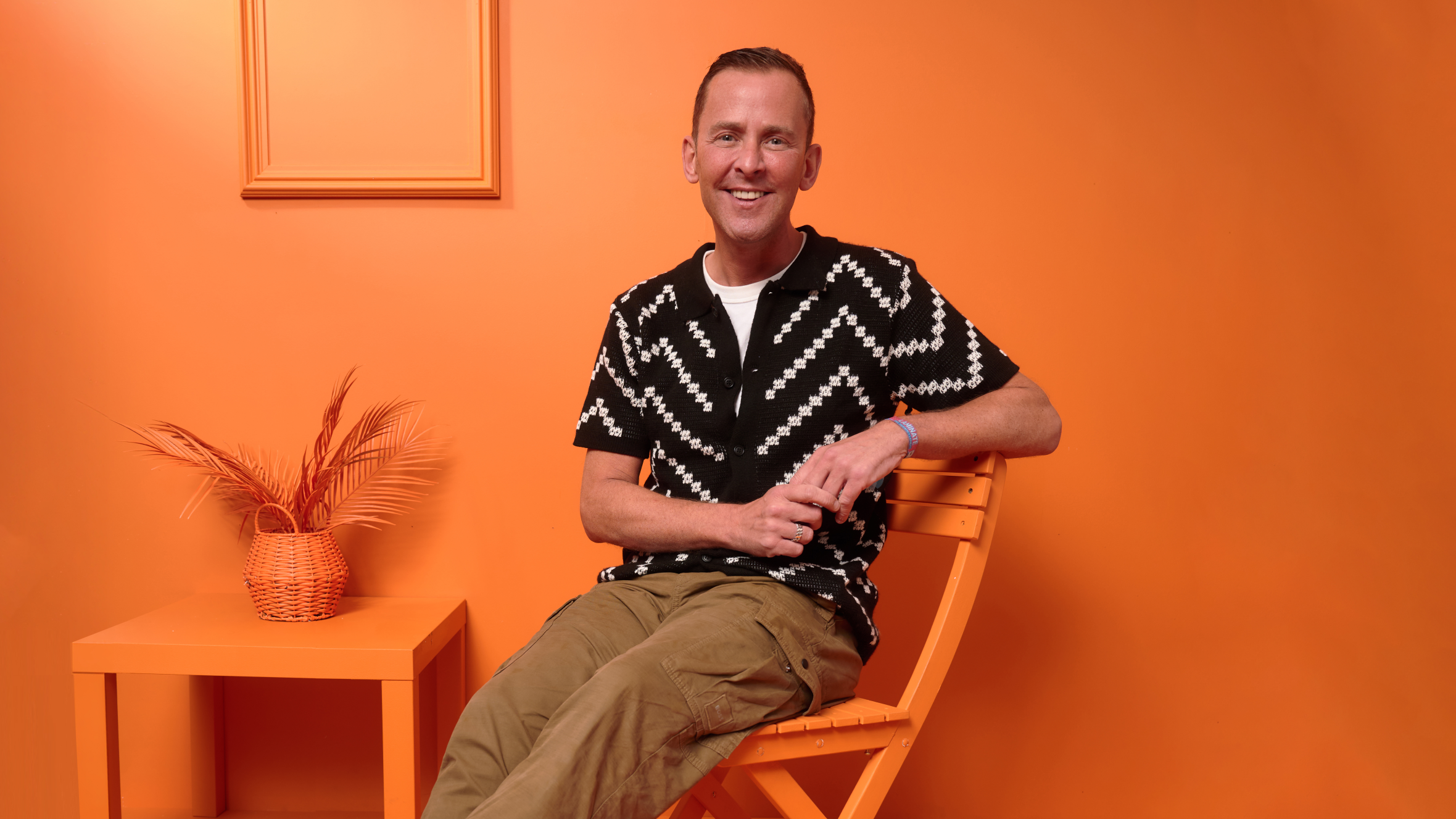 Scott Mills, in a black and white patterned shirt, smiling while seated in an orange room.