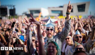 Eurovision fans watch the 2023 Eurovision Song Contest final on a giant screen in host city Liverpool. Many fans are dressed up, including the man in the centre of the photo who wears a silver jumpsuit and sunglasses and wears a silver cap with a giant silver star on the top.