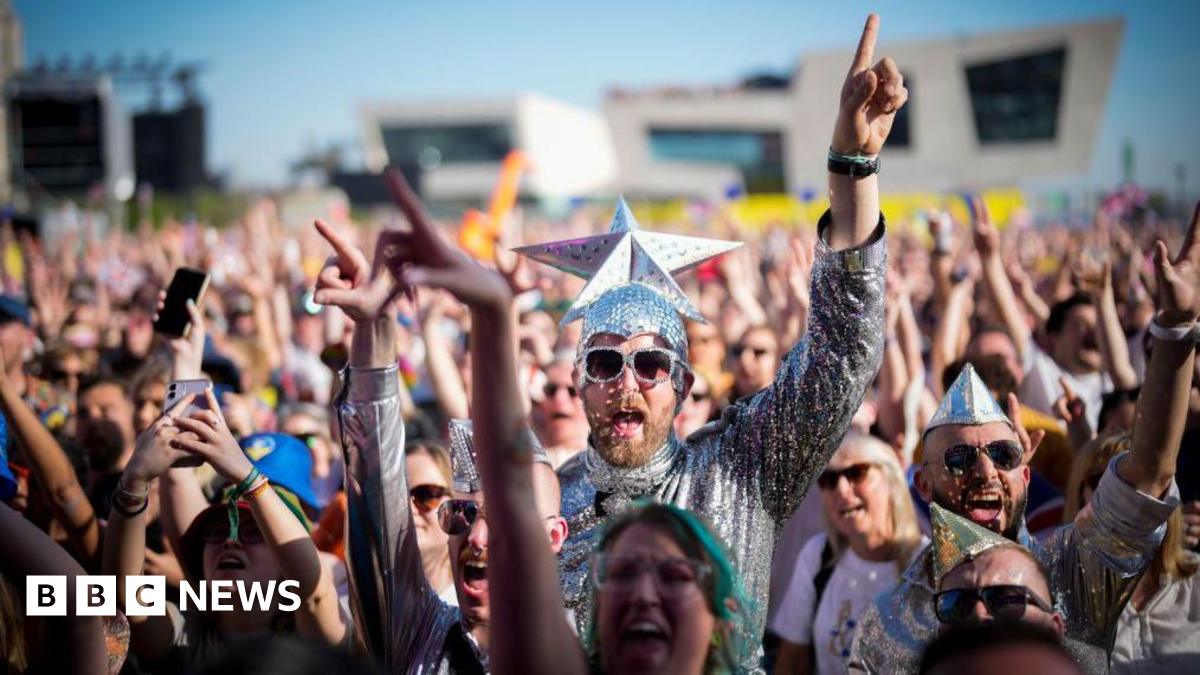 Eurovision fans watch the 2023 Eurovision Song Contest final on a giant screen in host city Liverpool. Many fans are dressed up, including the man in the centre of the photo who wears a silver jumpsuit and sunglasses and wears a silver cap with a giant silver star on the top.
