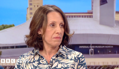 Shirine Khoury-Haq with short brown hair, earing a blue shirt with a white leaf print on it, sitting on the BBC Breakfast sofa with the Imperial War Museum North seen behind her