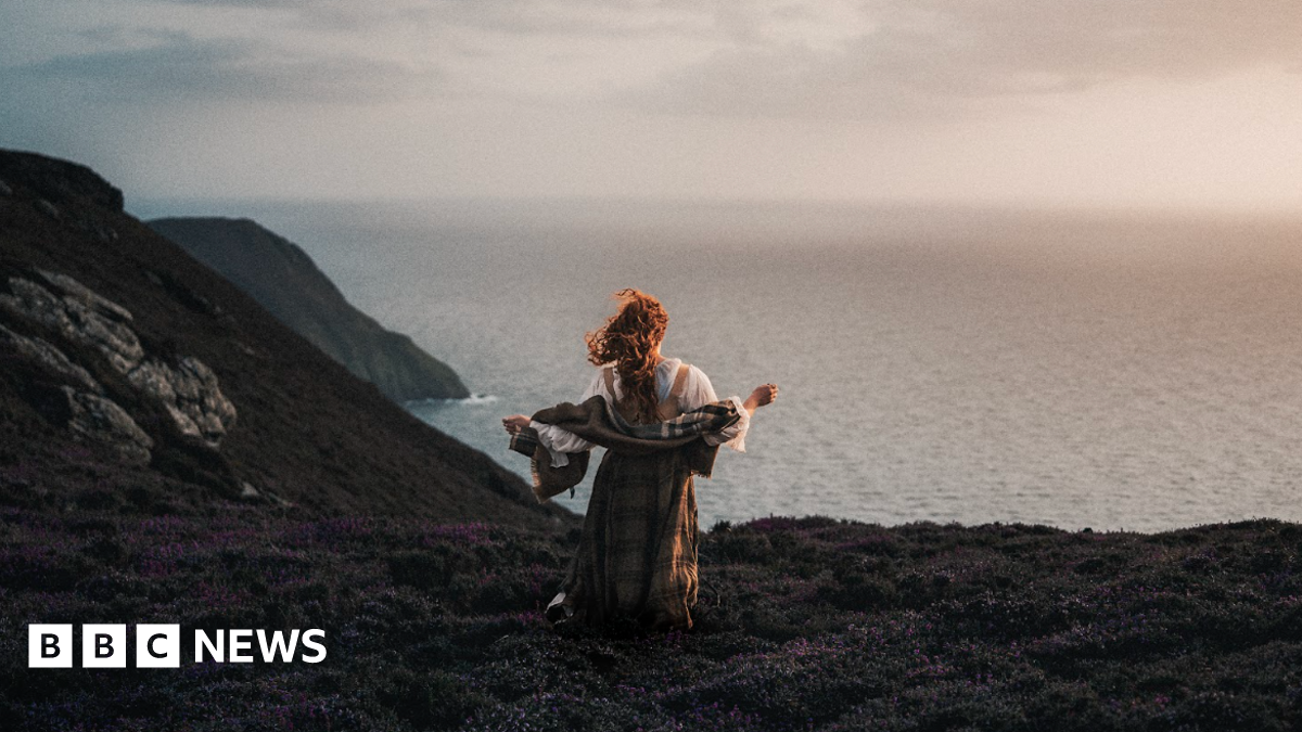 Ciara stands wearing traditional Celtic clothing on the edge of a cliff looking out to sea.