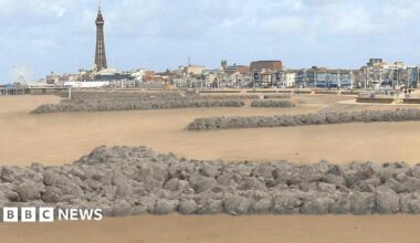 Visualisation of rock headlands along the beach. They are long stretches of grey rocks edging from the steps to the beach towards the sea. Blackpool's coastline can be seen in the distance in the background with the tower to the left